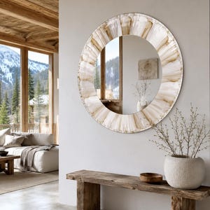 May include: A round wall mirror featuring a wide, textured frame with a radial pattern of translucent, cream and beige glass segments. The mirror surface reflects a room interior, while the frame displays a distinct, layered, fan-like design.