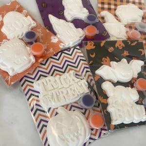 Op de afbeelding: Halloween knutselsets met witte gipsfiguren zoals geesten en pompoenen om te schilderen. Elke set bevat kleine potjes oranje en paarse verf. De sets zijn verpakt in transparante zakken met patronen op de achtergrond.