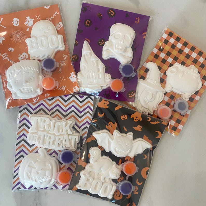 Op de afbeelding: Een verzameling Halloween knutselpakketten. Elk pakket bevat een wit gipsfiguur, zoals een spook of pompoen, met verfpotjes in oranje en paars. De pakketten zijn verpakt in decoratieve zakjes met Halloween patronen.