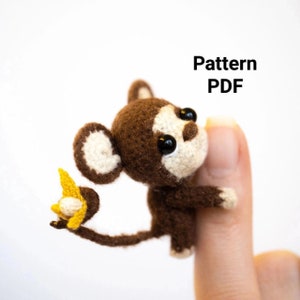 Puede incluir: Un mono de ganchillo con pelaje marrón, cara y orejas color crema y ojos negros. Sostiene un plátano amarillo. El texto "Patrón PDF" se muestra en letras negras.