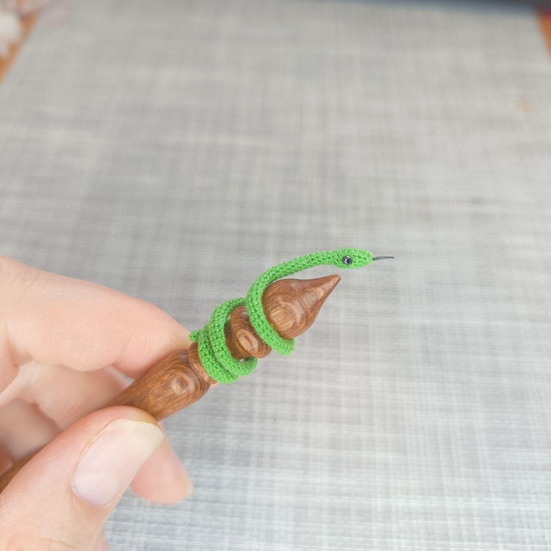 K&ouml;nnte beinhalten: Eine gr&uuml;ne geh&auml;kelte Schlange, die um eine h&ouml;lzerne Stricknadel gewickelt ist.
