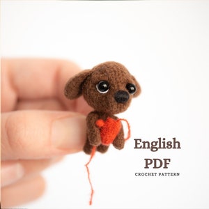 Op de afbeelding: Een bruine gehaakte hond die een rood hart vasthoudt. De hond heeft zwarte ogen en een zwarte neus. De tekst "English PDF Crochet Pattern" staat onder de hond.