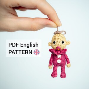 Może przedstawiać: Przędza amigurumi z ubraniem w kolorze różowym i białym, smoczkiem i aureolą. Lalka jest trzymana przez rękę. PDF English PATTERN.