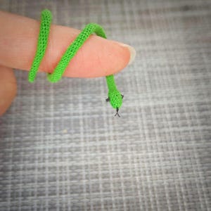 K&ouml;nnte beinhalten: Ein gr&uuml;nes geh&auml;keltes Schlangenspielzeug, das um einen Finger gewickelt ist.