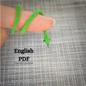 K&ouml;nnte beinhalten: Eine gr&uuml;ne geh&auml;kelte Schlange, die sich um einen Finger windet. Die Schlange ist klein und detailliert, mit einer schwarzen Perle als Auge. Der Text "English PDF Crochet Pattern" befindet sich unter der Schlange.