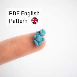 Könnte beinhalten: Ein kleines blaues Häkel-Nilpferd-Spielzeug, das auf einem Finger sitzt. Das Spielzeug hat schwarze Augen und einen hellblauen Körper. Das Bild enthält den Text "PDF English Pattern" und ein kleines Bild der britischen Flagge.