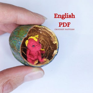 Op de afbeelding: Een kleine rode gehaakte draak met gele hoorns zit in een groen-gouden eivormige container. De tekst "English PDF CROCHET PATTERN" staat in rode letters boven het ei.