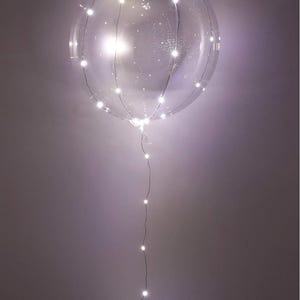 Puede incluir: Un globo transparente y redondo iluminado con pequeñas luces LED blancas brillantes. Las luces están dispuestas alrededor de la superficie del globo y se extienden hacia abajo por un cable delgado. El globo está suspendido sobre un fondo neutro.