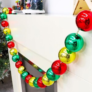 Puede incluir: Una colorida guirnalda navideña con adornos rojos, verdes y amarillos. Las brillantes bolas redondas están ensartadas, creando una decoración festiva. La guirnalda está colgada sobre una superficie blanca, cerca de un árbol de Navidad.