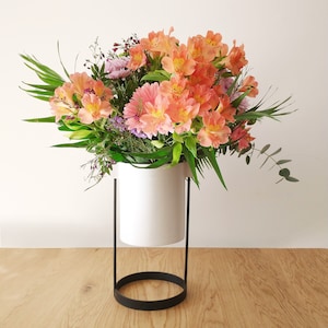 May include: A vibrant floral arrangement featuring peach-colored alstroemeria, pink gerbera daisies, and various greenery. The flowers are displayed in a white cylindrical vase with a black metal stand, set against a light wood surface.