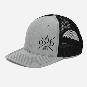 以下が含まれることがあります： グレーとブラックのトラッカーハット。ブラックの刺繍ロゴには、十字架が描かれた「DAD」と、その下に「EST 2025」と書いてあります。