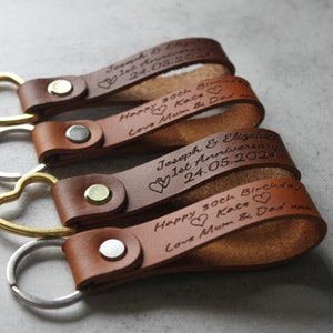 May include: Close-up of several brown leather keyrings with engraved text. The keyrings have heart-shaped and round metal attachments. The text includes names, dates, and messages, secured with metal rivets.