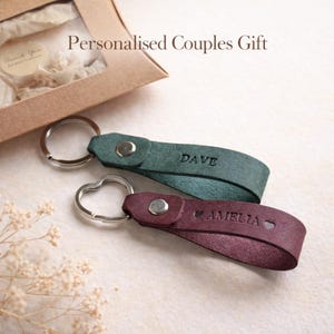 May include: Two personalised leather keyrings, one teal and one burgundy, with silver keyrings. The teal keyring is engraved with "DAVE", and the burgundy keyring is engraved with "AMELIA" and a heart symbol. The text "Personalised Couples Gift" is at the top.