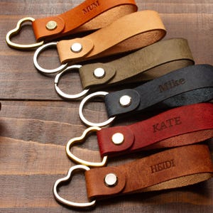 May include: A selection of leather keyrings in shades of brown, tan, green, navy, and red. Each keyring has a heart-shaped clasp and is engraved with a name or word, including "MUM," "Mike," "KATE," and "HEIDI."