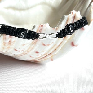 May include: Black friendship bracelet with an infinity symbol charm and black beads with the letters "S" and "L" on them.
