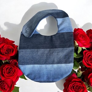May include: A denim tote bag with a curved handle and a button closure. The bag is made from horizontal panels of different shades of blue denim. Red roses are arranged around the bag.