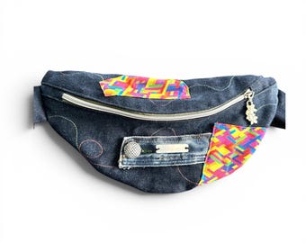 May include: A dark blue denim fanny pack featuring a silver zipper with a flower-shaped charm, decorative wavy stitching, and two patches of vibrant, multi-colored geometric fabric. A small denim pocket with a silver button and metal tag is attached.