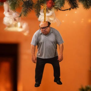 May include: A Christmas tree ornament featuring a figure of a man wearing glasses, a gray polo shirt, and black pants. The ornament is hanging from a Christmas tree branch. The background includes a fireplace and a decorated Christmas tree.