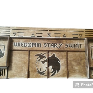 Puede incluir: Un organizador de juegos de madera con las palabras "Wiedźmin Stary Świat" y un símbolo de lobo. El organizador tiene compartimentos para piezas de juego y cartas.