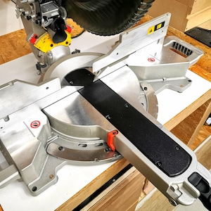 Puede incluir: Una sierra de inglete plateada y negra con una hoja negra y una palanca roja. La sierra está montada en una mesa de madera y tiene un transportador con marcas en grados.