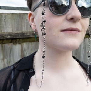 May include: A silver chain with black beads hangs from a pair of sunglasses. The chain is worn around the neck and has a small snake charm. The person wearing the chain has green hair and is wearing a black shirt.
