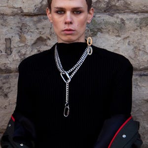 Op de afbeelding: Een persoon draagt een zwarte coltrui en een zilveren ketting met een karabijnhaaksluiting. De persoon draagt ook zilveren oorbellen en een zwarte jas met rode bies. De achtergrond is een stenen muur.