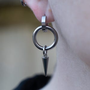 Op de afbeelding: Een zilveren oorring met een hangende zilveren punt. De oorring wordt in de oorlel gedragen.