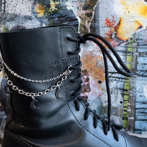 Puede incluir: Bota de cuero negro con cordones negros y un accesorio de cadena plateada. La cadena presenta peque&ntilde;os dijes de media luna. La bota se encuentra sobre un fondo abstracto colorido.