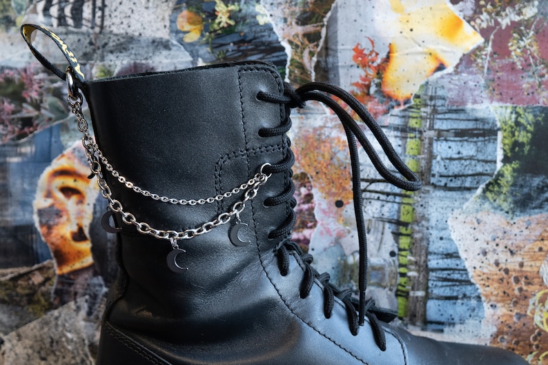 Puede incluir: Bota de cuero negro con cordones negros y un accesorio de cadena plateada. La cadena presenta peque&ntilde;os dijes de media luna. La bota se encuentra sobre un fondo abstracto colorido.