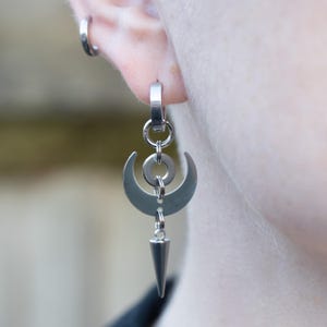 Puede incluir: Pendientes de aro de plata con un diseño de luna creciente, un círculo y una punta puntiaguda.