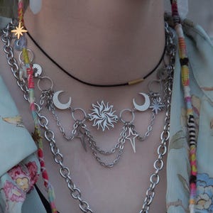 May include: A layered silver necklace featuring celestial charms. The necklace includes a sun pendant, crescent moons, and stars, with multiple chain strands. A black cord necklace and a gold star charm are also visible.