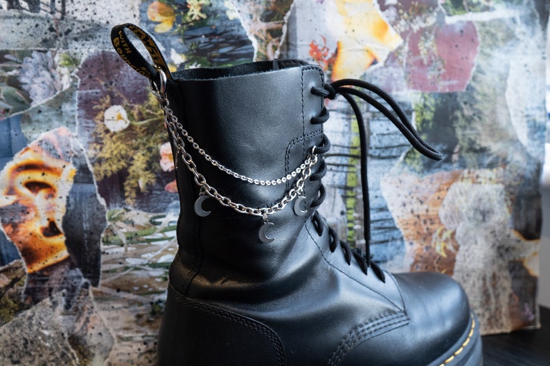 Puede incluir: Bota de combate de cuero negro con una cadena plateada y dijes de luna creciente. La bota tiene cordones negros y una suela cosida amarilla. El fondo es un collage de papel rasgado con elementos florales y abstractos.