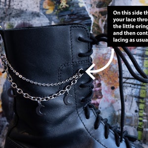 Puede incluir: Bota de cuero negro con cadena plateada y colgantes de luna creciente. La bota tiene cordones negros y un lazo en la parte superior. El texto de la imagen dice: "En este lado, enhebra tu cord&oacute;n a trav&eacute;s del peque&ntilde;o ojal y luego contin&uacute;a atando como de costumbre."