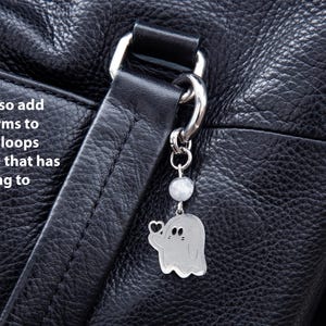 K&ouml;nnte beinhalten: Ein silberner Geisteranh&auml;nger mit Herzdetail h&auml;ngt an einem silbernen Verschluss an einer schwarzen Ledertasche. Der Anh&auml;nger ist an einer kleinen, runden, wei&szlig;en Perle befestigt. Der Text auf dem Bild lautet: "Sie k&ouml;nnen diese Anh&auml;nger auch an Taschen, G&uuml;rtelschlaufen oder allem, was eine Schlaufe/einen Ring zum Einh&auml;ngen hat, anbringen."