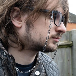 Puede incluir: Una persona lleva gafas con montura negra con una cadena plateada unida a las patillas. La cadena cuelga por la parte delantera del pecho de la persona.