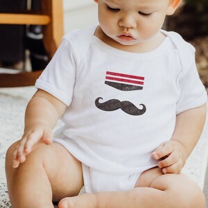 Può includere: Un body bianco per neonato con un baffo nero e strisce rosse e bianche sul davanti. Il bambino è seduto su un tappeto bianco.