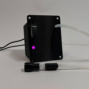 May include: A black carbon fiber patterned control box featuring a rectangular faceplate with four corner screw holes, a black toggle switch, and a glowing pink LED indicator light. Two clear plastic tubes extend from the unit, with one tube connected to a black cylindrical nozzle component.