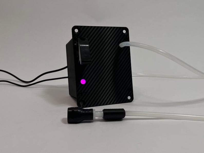 May include: A black carbon fiber patterned control box featuring a rectangular faceplate with four corner screw holes, a black toggle switch, and a glowing pink LED indicator light. Two clear plastic tubes extend from the unit, with one tube connected to a black cylindrical nozzle component.