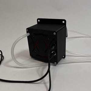 May include: A black rectangular electronic device with a carbon fiber textured front panel featuring a red circular design. The unit has a mounting flange, two clear flexible plastic tubes extending from the sides, and two black power cables.