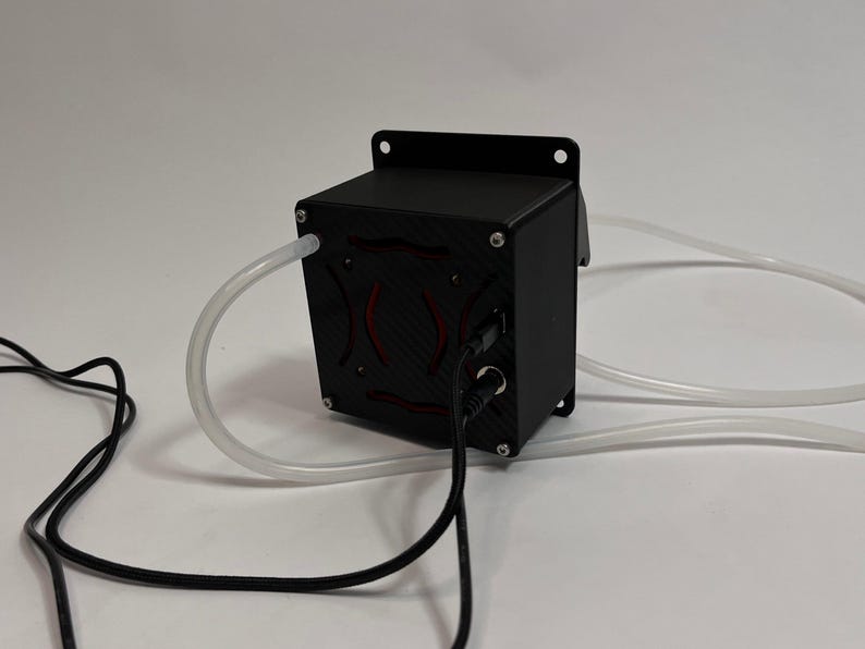 May include: A black rectangular electronic device with a carbon fiber textured front panel featuring a red circular design. The unit has a mounting flange, two clear flexible plastic tubes extending from the sides, and two black power cables.