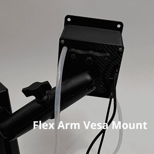 May include: A black Flex Arm Vesa Mount featuring a rectangular housing with a carbon fiber pattern, a central adjustment knob, and a vertical support post. The device includes a clear plastic tube and black electrical cables extending from the unit.