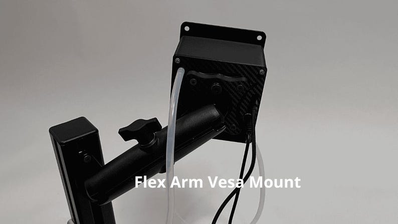 May include: A black Flex Arm Vesa Mount featuring a rectangular housing with a carbon fiber pattern, a central adjustment knob, and a vertical support post. The device includes a clear plastic tube and black electrical cables extending from the unit.