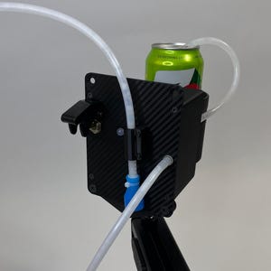 May include: A black device with a carbon fiber texture holds a green can. Clear tubing connects to the can and the device. The device has a black bracket and a blue connector. The background is a plain white.