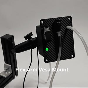 May include: A black Flex Arm Vesa Mount featuring a carbon fiber patterned rectangular plate with a small glowing green light, a black adjustable arm, and two clear plastic tubes extending from the front, all set against a plain background.