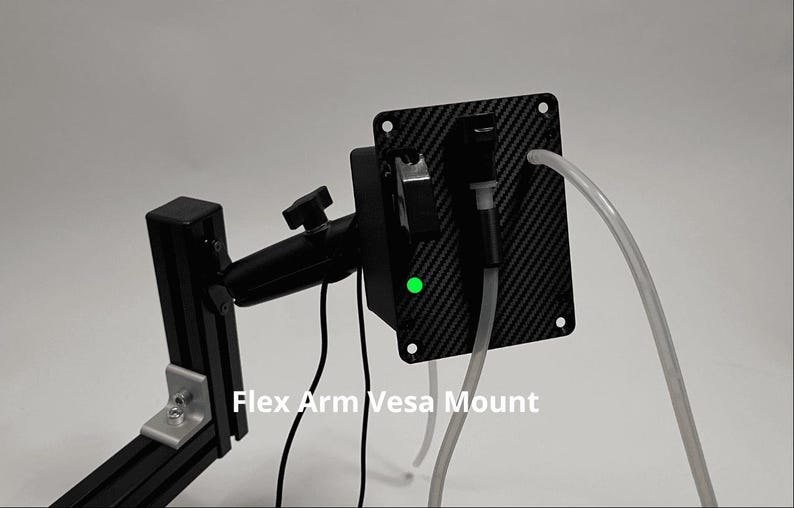 May include: A black Flex Arm Vesa Mount featuring a carbon fiber patterned rectangular plate with a small glowing green light, a black adjustable arm, and two clear plastic tubes extending from the front, all set against a plain background.