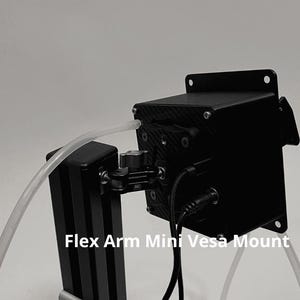 May include: A black Flex Arm Mini Vesa Mount featuring a square housing with a carbon fiber pattern, a central adjustable joint, and a vertical support column. A clear plastic tube and black power cables are connected to the device.