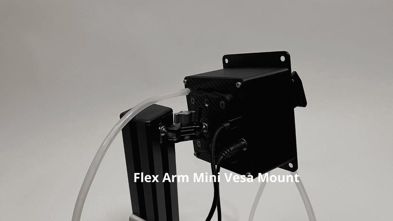 May include: A black Flex Arm Mini Vesa Mount featuring a square housing with a carbon fiber pattern, a central adjustable joint, and a vertical support column. A clear plastic tube and black power cables are connected to the device.