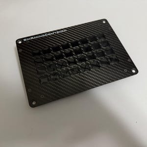May include: A carbon fiber sim racing button box with multiple black buttons. The text "SIMRACINGCRAFTSMEN" is printed in white on the top left corner. The button box has a rectangular shape with rounded corners and mounting holes.