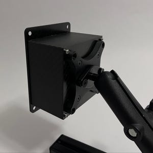 Könnte beinhalten: Ein schwarzes, rechteckiges Gerät mit strukturierter Oberfläche, montiert auf einem schwarzen, verstellbaren Arm. Das Gerät hat eine quadratische Form mit abgerundeten Ecken und ist mit Schrauben befestigt. Der Arm hat mehrere Gelenke für flexible Positionierung.