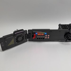 Puede incluir: Un dispositivo electrónico negro con la palabra "HELLO" en rojo. El dispositivo tiene múltiples botones, luces y un ventilador. El dispositivo está abierto, revelando componentes internos. El dispositivo está etiquetado "SIMRACINGCRAFTERS".
